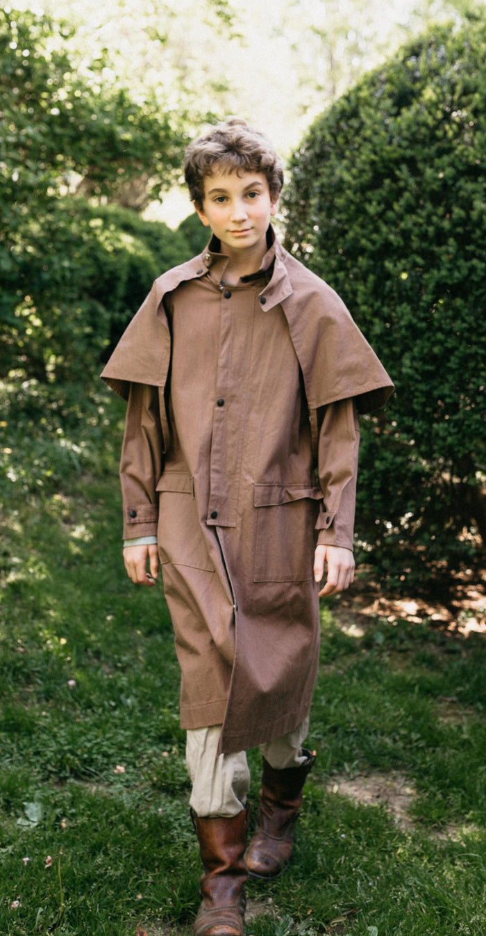 Older boy walking toward camera wearing a brown canvas Drover's Coat. Outside in front of green bushes.