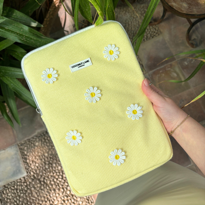 Hand holding a yellow iPad sleeve with white daisy pattern by Coconut Lane, surrounded by green plants.
