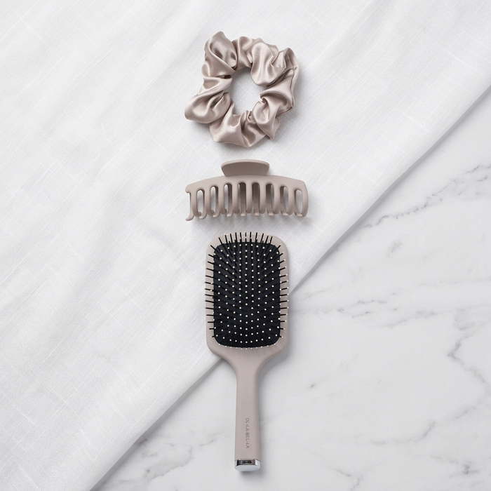 Three haircare accessories, the Olla Bella Everyday Glam Hair Accessory Kit, arranged vertically on a white textured fabric over a white marble surface: a satin scrunchie, a claw comb, and a paddle brush. The brand name Olla Bella is subtly printed near the base of the brush handle.