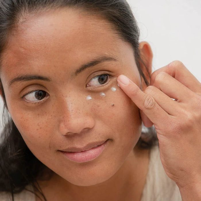 Woman eye balm to her face with a neutral background.