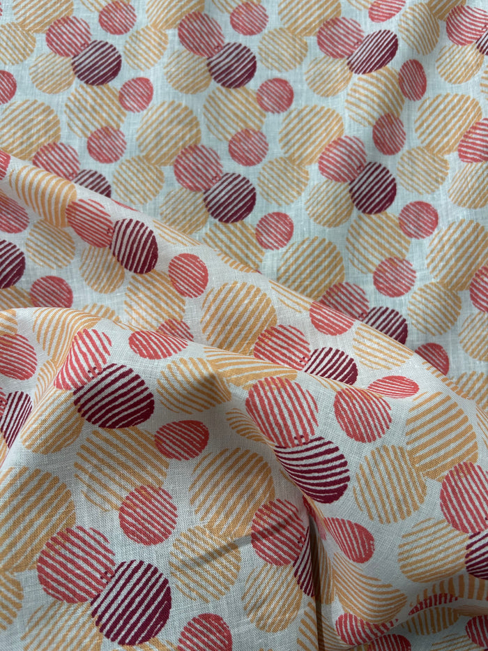 Italian linen with a retro abstract polka dot print fabric in orange, red, and burgundy on an ivory background. The circles vary in size and are filled with fine diagonal stripe textures, creating a layered geometric look with a hand-printed feel.
