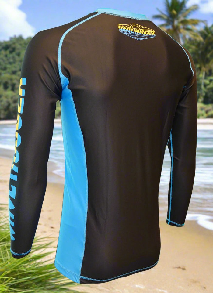 The Wave Hugger Blue Panel Rash Guard features black and blue fast-drying lycra, built-in sun protection, and WAVE HUGGER on the sleeve and upper back, shown on a beach with palm trees, ocean waves, and greenery.