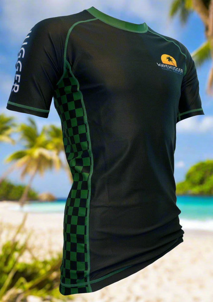 The Wave Hugger Classy and Nasty Rash Guard, in black and green with checkered side panels and a sunset logo, is displayed on a tropical beach with palm trees, capturing jiu jitsu spirit under a blue sky.