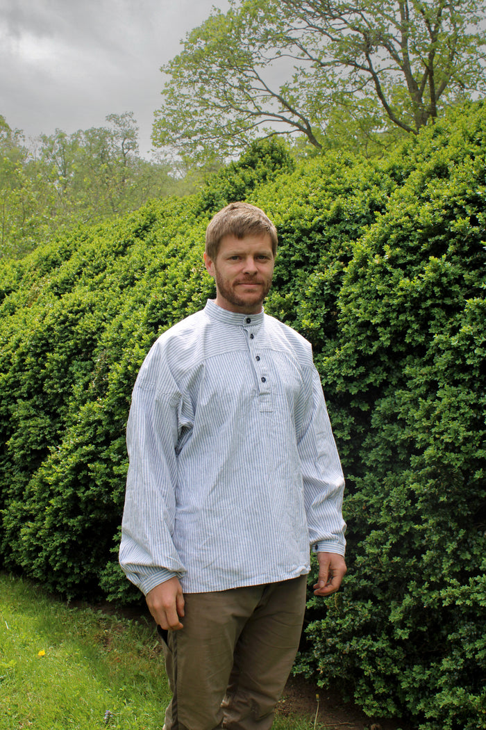 Man wearing a light-colored blue and white striped shirt standing in front of green bushes