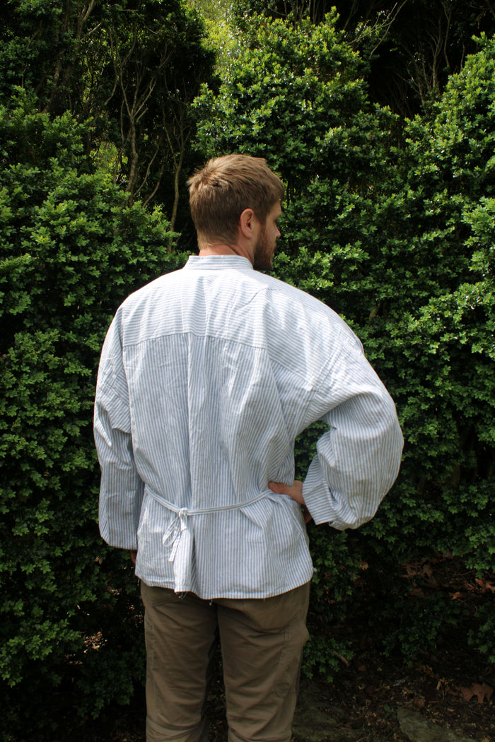 Back of a man with his right hand on his hip wearing a blue and white striped shirt standing in front of green bush.