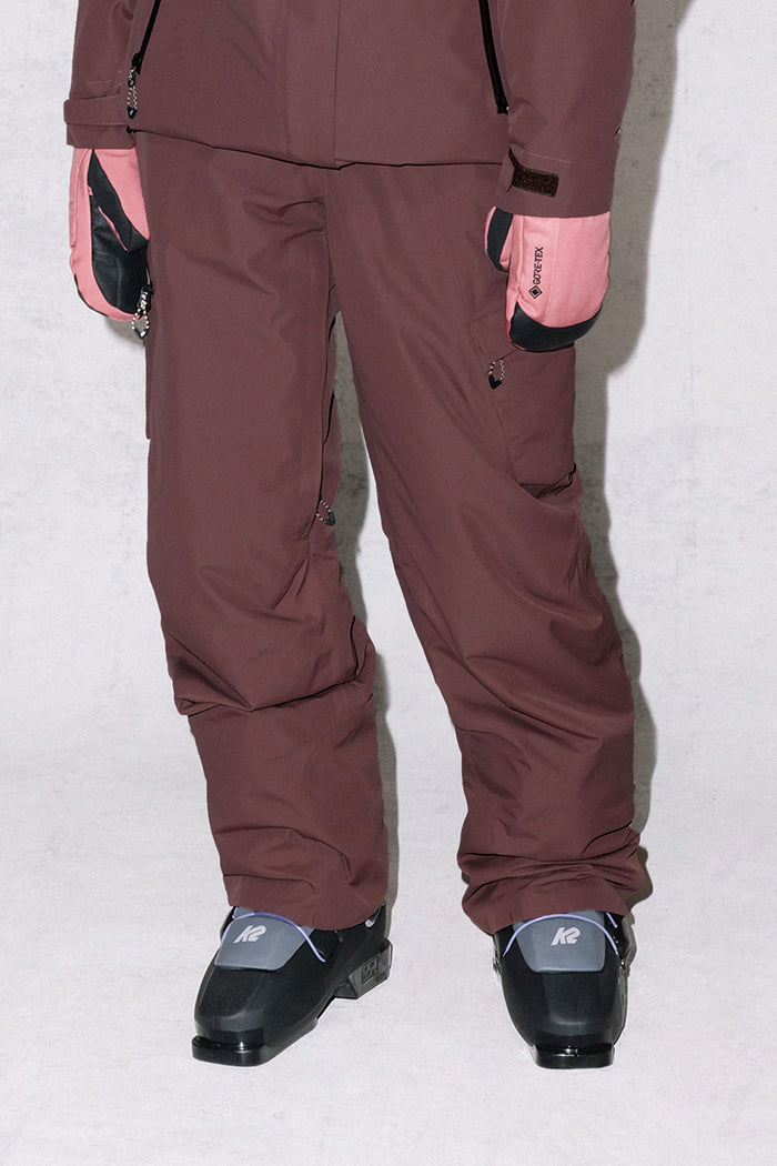 A person wearing 686 Women's Geode Thermagraph® Pant in brown, a matching jacket, pale pink gloves, and black K2 ski boots stands against a light background. Only the lower body is visible.