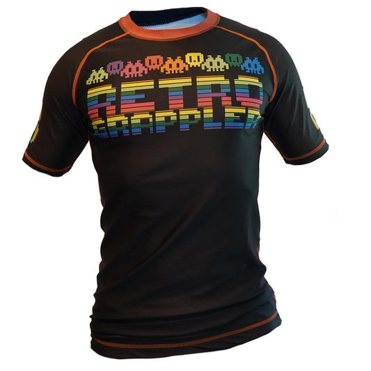 The 8-Bit Rainbow Rash Guard by Retro Grappler is a black short-sleeved BJJ top with vibrant pixelated space invader graphics and RETRO GRAPPLER in rainbow arcade style—ideal for no-gi sessions.
