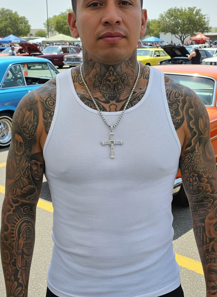 Man with tattoos wearing a white tank top and cross necklace in a car show setting