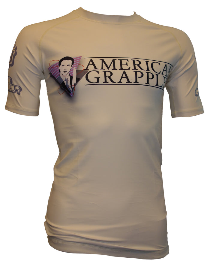 The Retro Grappler American Grappler Rash Guard is beige, features AMERICAN GRAPPLER across the chest, a man in a suit with purple wings on the left side, and small MMA logos on both sleeves.