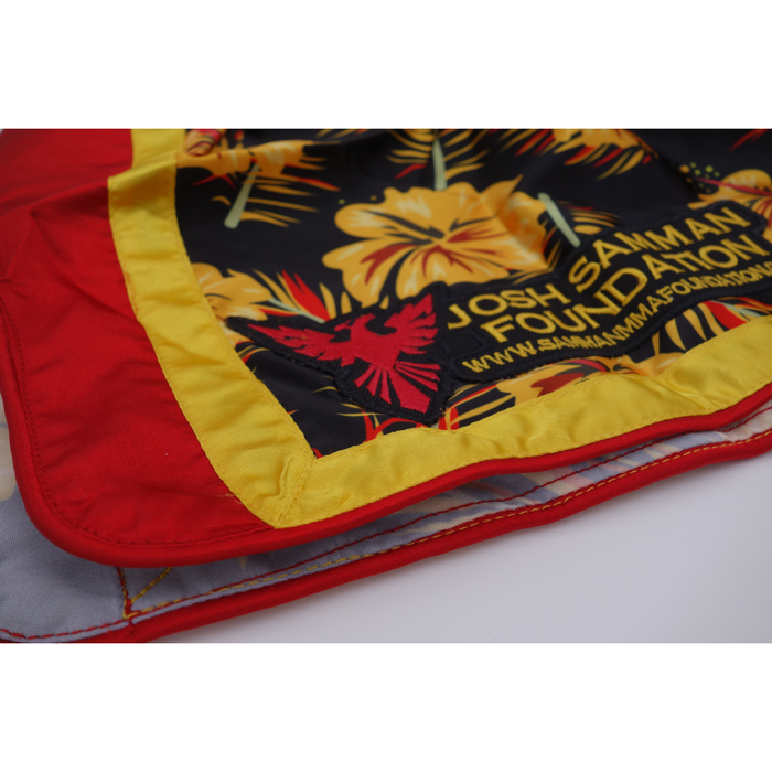 A close-up of Combat Nights Black Gold Red Hawaiian Style Muay Thai shorts shows colorful floral patterns, a yellow border, red backing, the Josh Samman Foundation text, a red bird logo, and the website URL.