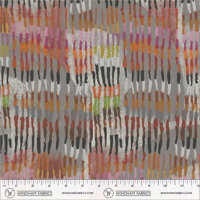 A close-up of a fabric pattern by Windham Fabrics featuring vertical brushstroke-like dabs in pink, orange, yellow, green, black, brown, and white over a mottled gray background, with a printed ruler scale along the bottom edge.