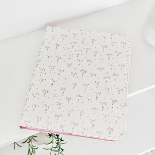 Coconut Lane iPad case with pink bow pattern, displayed on a white shelf next to a green plant.