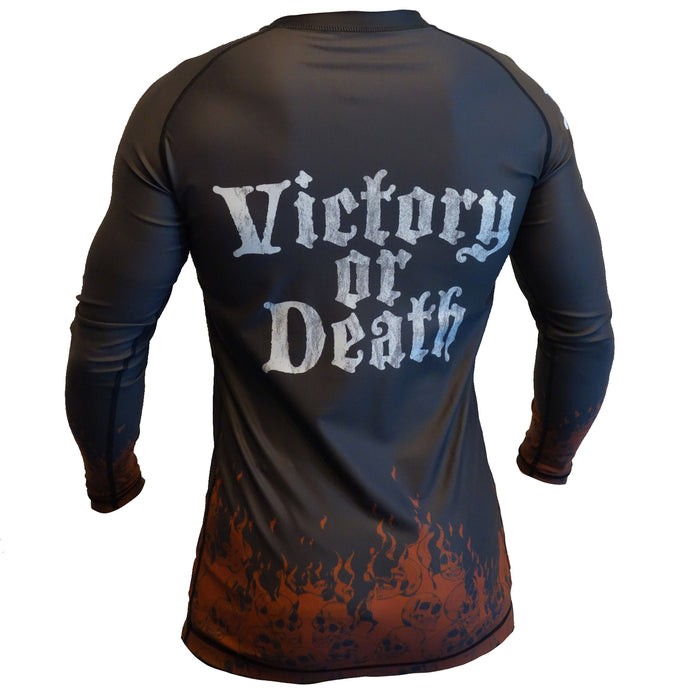 A person wearing the Retro Grappler Death Before Submission Motorhead Jiu Jitsu Rash Guard, featuring Victory or Death in bold white letters on the back and a brown flame and skull pattern along the bottom.