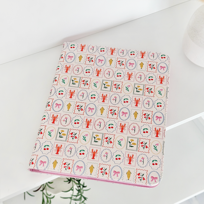Dolce Vita iPad case with a colorful pattern on a white shelf next to a patterned notebook and small green plant.