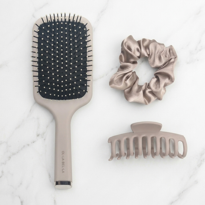The Olla Bella Everyday Glam Hair Accessory Kit featuring a paddle hairbrush, a satin scrunchie, and a claw clip, all in a matching taupe color, arranged on a white marble background.
