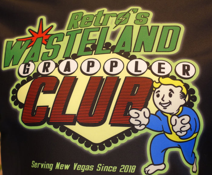 A colorful graphic reads Fallout Wasteland Grappler Club with a cartoon character giving a thumbs-up and the tagline Serving New Vegas Since 2018, inspired by our Retro Grappler limited edition rash guard.
