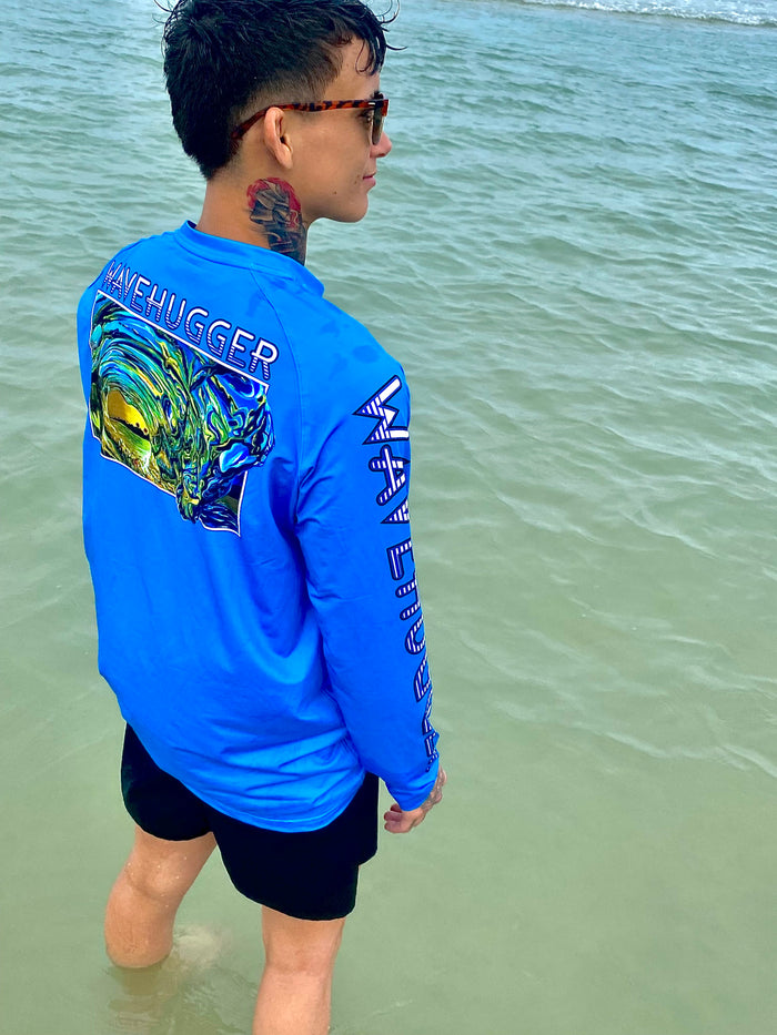 A person in a bright blue Wave Hugger Hawaiian Gold Performance Shirt and black shorts stands in shallow ocean water, looking away. The moisture-wicking shirt offers UPF 50+ sun protection and a colorful wave design on the back.