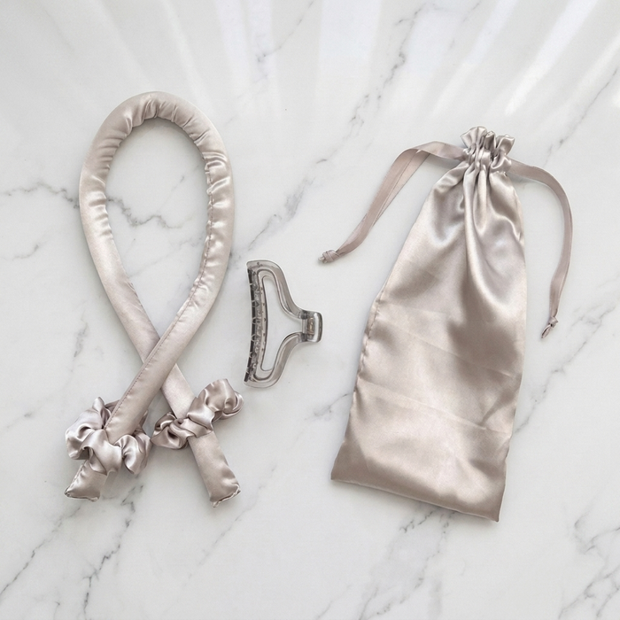 A satin hair accessory set laid out on a white marble surface, including the Olla Bella Satin Heatless Curling Headband, two satin scrunchies, a claw clip, and a satin drawstring bag.