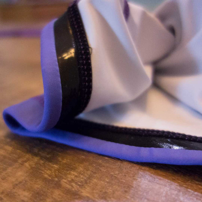 A close-up of the Retro Grappler Heel Hookers Ball Heel Hook Rash Guard, featuring white fabric with purple trim and black piping, resting on a wooden surface.