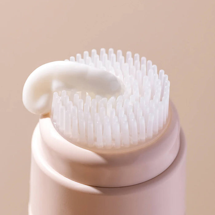 Close-up of a pink facial cleansing brush with bristles and a white handle on a beige background.