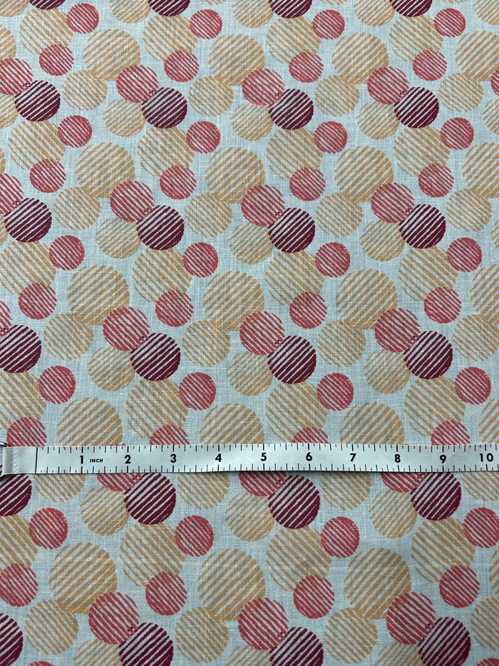 Italian linen with a retro abstract polka dot print fabric in orange, red, and burgundy on an ivory background. The circles vary in size and are filled with fine diagonal stripe textures, creating a layered geometric look with a hand-printed feel next to a ruler showing scale.