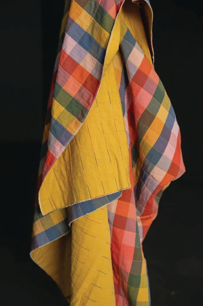 Colorful checked print of yellow, oranges, blues, purples, and pinks  draped over a yellow garment on a black background