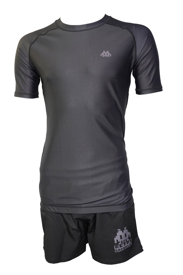 The Retro Grappler Just a black rash guard and shorts set features a fitted, short-sleeve shirt and matching grappling shorts, each with a small pixelated light gray logo on the chest and left leg. Shown on a headless mannequin.