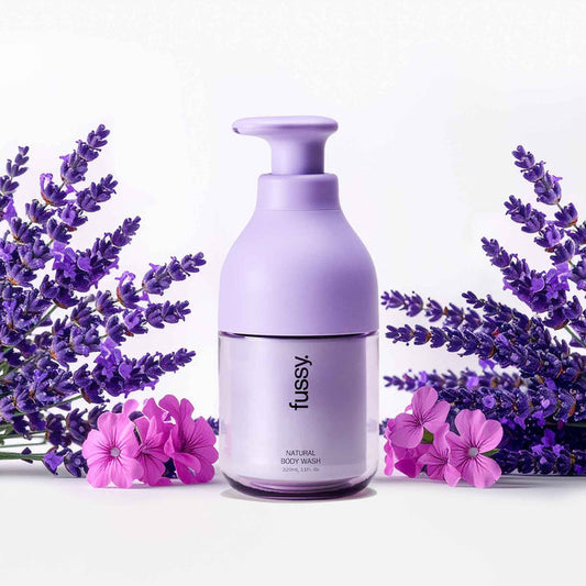 Fussy's natural, refillable Lavender Fields body wash in a Lilac pump bottle, set against a background of lavender and geranium flowers.