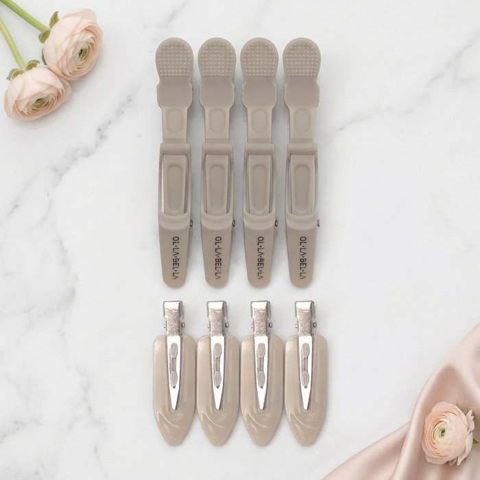Four Olla Bella Crocodile Clips and four Olla Bella Creaseless Clips from our Olla Bella Mane Master Hair Clip Set arranged in two rows of two on a light marble background with blush-pink flowers and a subtle piece of blush satin fabric.