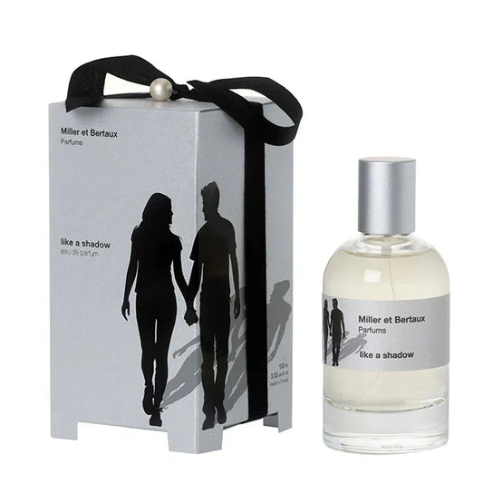 Miller et Bertaux Perfums 'like a shadow' eau de parfum, featuring a silver-gray gift box with a black ribbon and silhouette graphic next to a clear glass bottle with a silver cap.