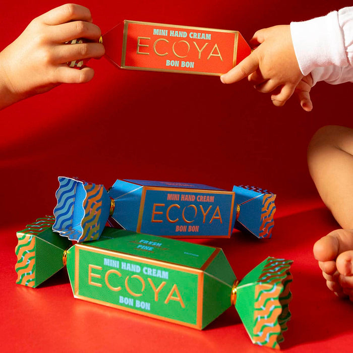 ECOYA mini hand cream boxes on a red background with hands interacting with one of the boxes.