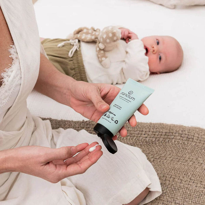 Person applying a skincare product to their hand with a baby lying on a blanket in the background.