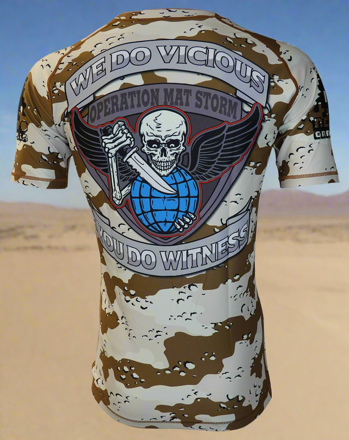 A person wears the Retro Grappler Operation Mat Storm Desert Camo Rash Guard, featuring a back patch with a winged skull holding a blue globe and the text WE DO VICIOUS, YOU DO WITNESS, OPERATION MAT STORM.