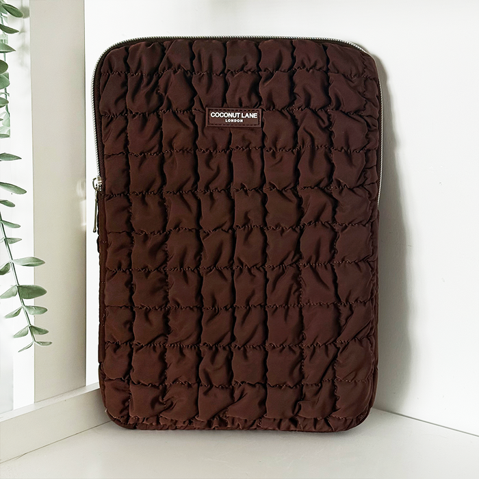 Quilted brown iPad sleeve with padded texture, placed on a white surface beside green leaves.