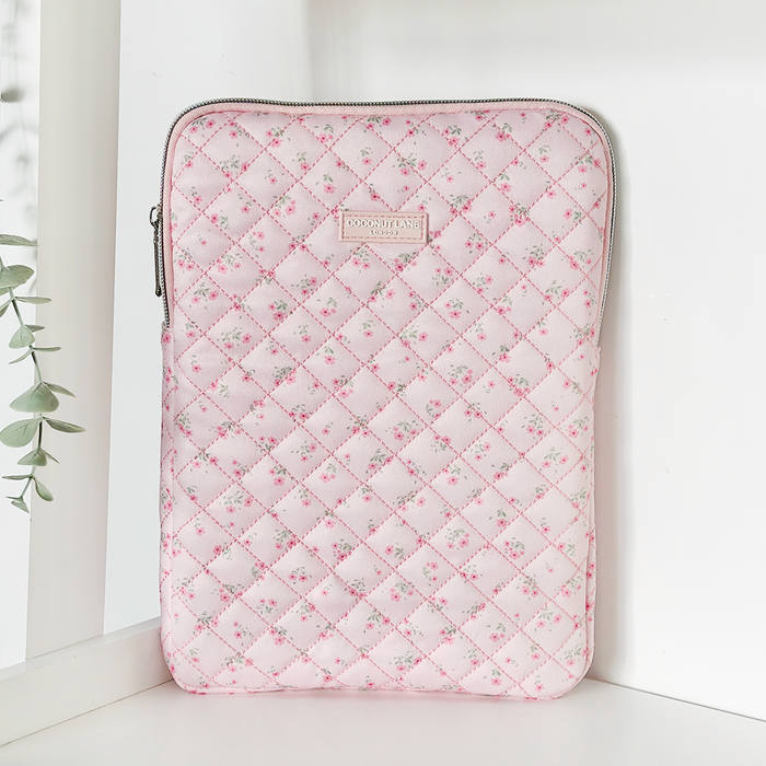 Pink quilted iPad sleeve with ditsy floral pattern and zipper, standing upright on a white surface near a plant.