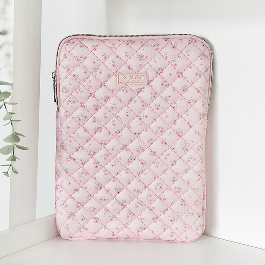 Pink quilted iPad sleeve with ditsy floral pattern and zipper, standing upright on a white surface near a plant.
