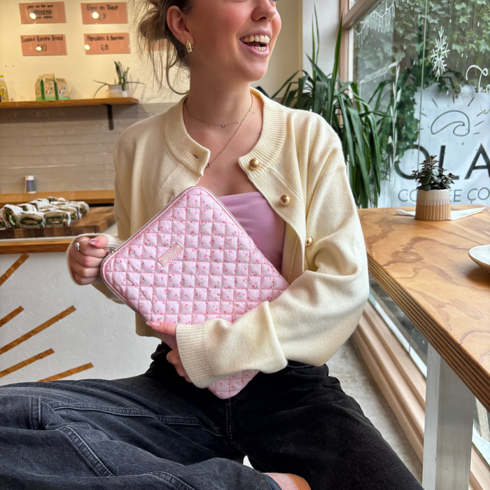 Woman holds a pink quilted iPad sleeve with watercolor flower pattern, standing by a sunny café window.
