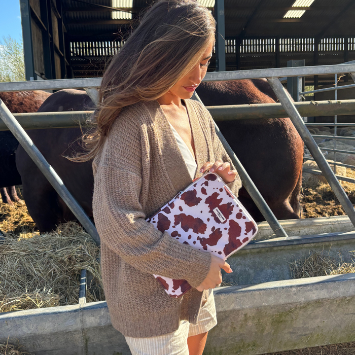 Woman holds a Rodeo Cowgirl iPad sleeve with a beige cardigan, standing in a sunlit barn by cows.