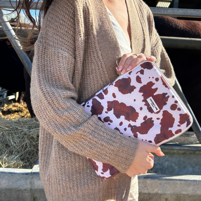 Woman in a tan cardigan holds a pink patterned Coconut Lane iPad sleeve, standing outdoors near cows.