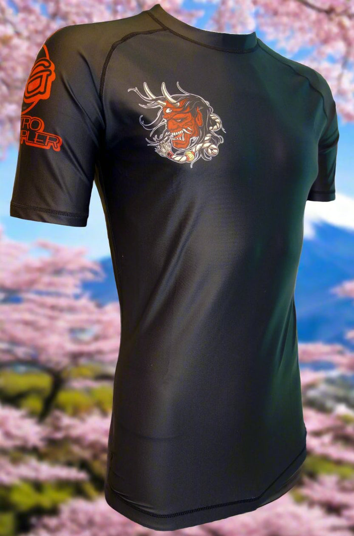 The Retro Grappler “Samurai Prodigy” Rash Guard is a black athletic shirt with Japanese Oni-inspired art on the chest and red logo text on the sleeve—ideal for jiu jitsu, shown against cherry blossoms and a blue sky.