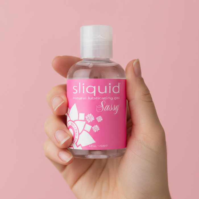 Handheld scale photo of the clear 4.2 fl oz (125 ml) bottle of Sliquid Sassy natural lubricating gel with its signature pink floral label held against a solid pink backdrop.