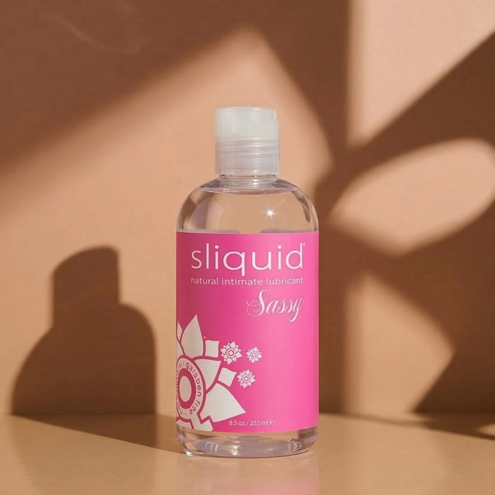 Front product shot of the clear 8.5 oz (255 ml) bottle of Sliquid Sassy natural intimate lubricant with a bright pink label, standing on a neutral surface casting warm leaf shadows.