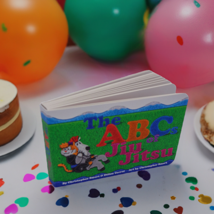 The ABCs of Jiu Jitsu Childrens Book by Retro Grappler sits on a table with confetti, cakes, and bright balloons in the background, creating a lively celebration of martial arts for kids.