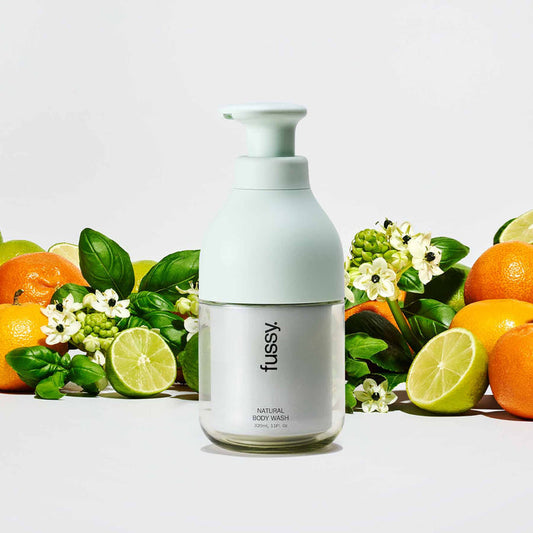 Fussy's Tropic Tonic natural body wash pump bottle, set against a background of limes, mandarins and basil leaves.