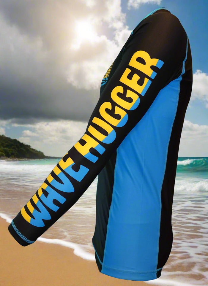 A person wears the Wave Hugger Blue Panel Rash Guard—long-sleeved and made from fast-drying lycra with bold yellow Wave Hugger branding—while standing on a sandy beach with ocean waves and a cloudy sky behind.