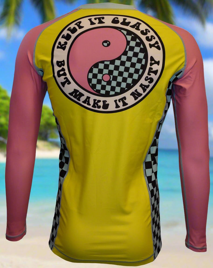 The Wave Hugger Classy and Nasty Rash Guard by Wave Hugger features yellow and pink long sleeves, a back yin-yang, checkered details, and “Keep it classy but make it nasty”—perfect surfwear for tropical beach vibes.