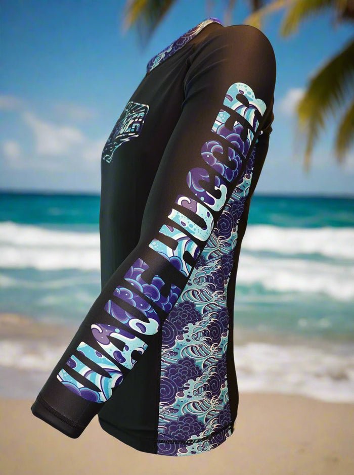 The Wave Hugger Japanese Wave Rash Guard by Wave Hugger is a unisex black lycra top with blue and purple wave patterns, BEACH ACCESS on the sleeve, offering fast-drying sun protection—shown beachside with sand, ocean waves, and palm trees.