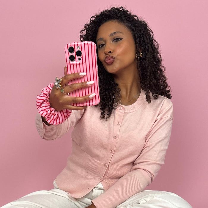 Curly-haired woman in pink holds a wavy phone case with red and white cherry stripes, taking a mirror selfie.