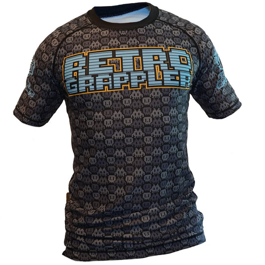 The Youth - 8-Bit Grey Rash Guard by Retro Grappler is a black short-sleeve BJJ top with a blue, orange, and white pixelated logo on the chest and an all-over gray pixel pattern. Affordable and perfect for young jiu jitsu athletes.