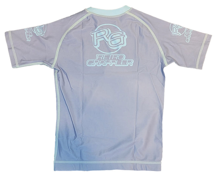 The Youth - Competition Shark by Retro Grappler is a light purple short-sleeve rash guard with white RG and Retro Grappler logos on the back and sleeves, light blue seam trim, four-way stretch fabric, and a fitted design shown laid flat (back view).